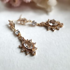 May include: A pair of gold-coloured drop earrings with clear, sparkling crystals. Each earring features a central, teardrop-shaped crystal surrounded by smaller crystals, with a delicate chain and additional crystals.