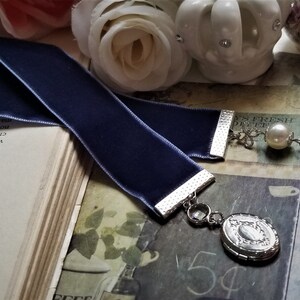 May include: A dark blue velvet bookmark with a silver metal clasp and a silver charm with a pearl and a crystal. The bookmark is lying on a book with text.