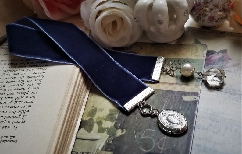 May include: A dark blue velvet bookmark with a silver metal clasp and a silver charm with a pearl and a crystal. The bookmark is lying on a book with text.