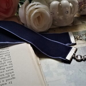 May include: A navy blue velvet bookmark with a silver chain and a silver locket charm. The locket has a white design inside. The bookmark is lying on a book with white pages.