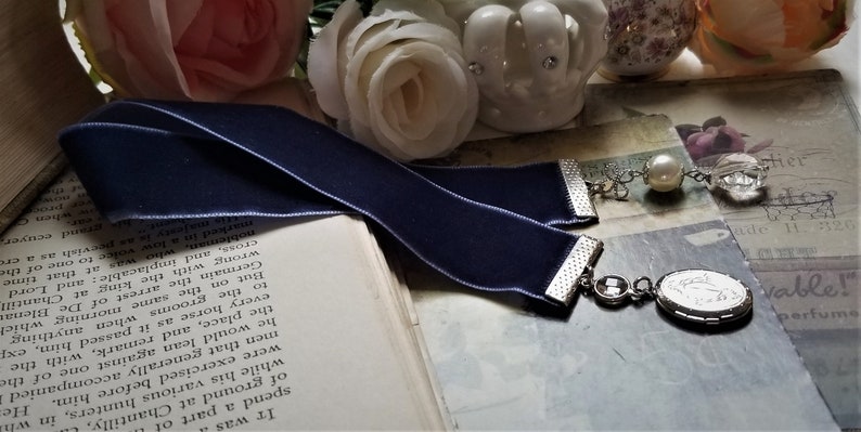 May include: A navy blue velvet bookmark with a silver chain and a silver locket charm. The locket has a white design inside. The bookmark is lying on a book with white pages.