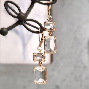 Peut inclure: Boucles d'oreilles dorées avec trois pierres de cristal transparentes rectangulaires. Les boucles d'oreilles sont suspendues à un crochet doré et sont présentées sur un présentoir décoratif en métal noir. Les cristaux ont une légère teinte champagne.