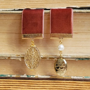 May include: A pair of earrings with a burgundy velvet ribbon and gold accents. One earring features a clear glass oval with a gold filigree design. The other earring features a gold locket with a heart design and a white pearl.