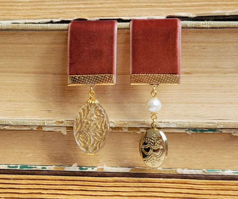 May include: A pair of earrings with a burgundy velvet ribbon and gold accents. One earring features a clear glass oval with a gold filigree design. The other earring features a gold locket with a heart design and a white pearl.