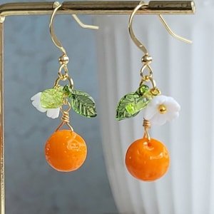 Puede incluir: Un par de pendientes colgantes de oro con una pequeña fruta naranja, hojas verdes y una flor blanca.