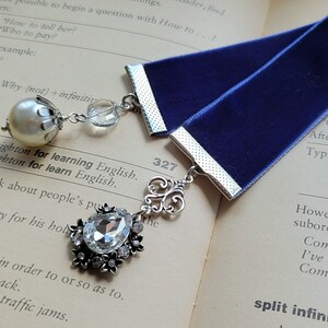 May include: A blue velvet bookmark with silver accents and two charms. One charm is a pearl with a silver crown and the other is a crystal with silver embellishments. The bookmark is laying on a book with the page number 327 visible.