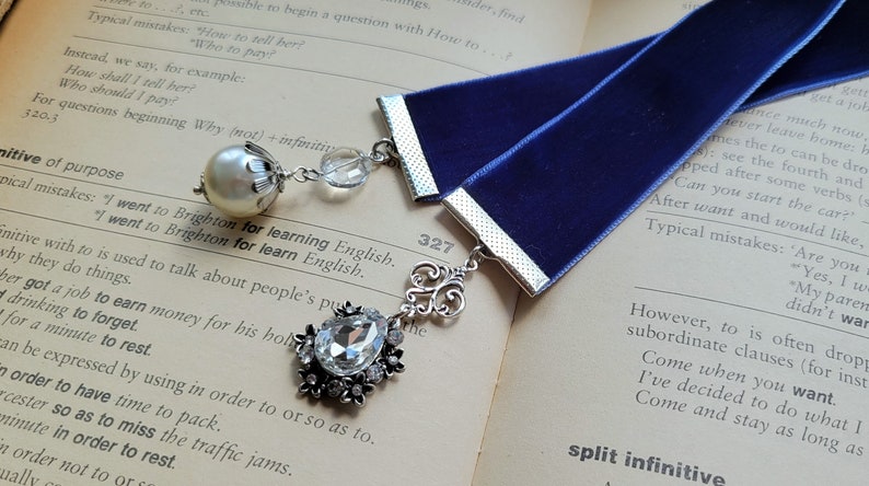 May include: A blue velvet bookmark with silver accents and two charms. One charm is a pearl with a silver crown and the other is a crystal with silver embellishments. The bookmark is laying on a book with the page number 327 visible.