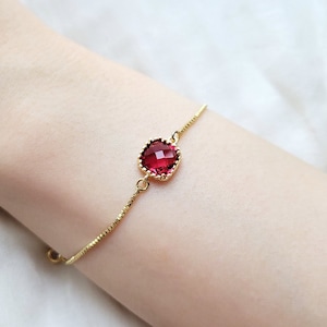 May include: A delicate gold chain bracelet with a single red gemstone pendant. The gemstone is faceted and set in a gold frame.