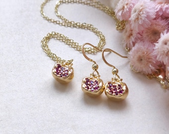 Può includere: Un set di gioielli color oro composto da una collana e un paio di orecchini. Ogni pezzo presenta un design a forma di melograno, con un gruppo di piccole gemme rosse. Il set è esposto su una superficie bianca, con fiori rosa sullo sfondo.