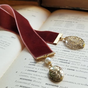 May include: Two burgundy velvet bookmarks with gold accents. One bookmark has a gold charm with a heart design and a pearl. The other bookmark has a clear glass charm with gold leaf accents. The bookmarks are on top of an open book.