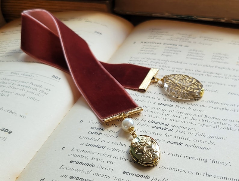 May include: Two burgundy velvet bookmarks with gold accents. One bookmark has a gold charm with a heart design and a pearl. The other bookmark has a clear glass charm with gold leaf accents. The bookmarks are on top of an open book.