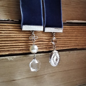 May include: Two blue velvet ribbon bookmarks with silver metal accents. One bookmark has a pearl and a crystal charm, the other has a silver locket charm.