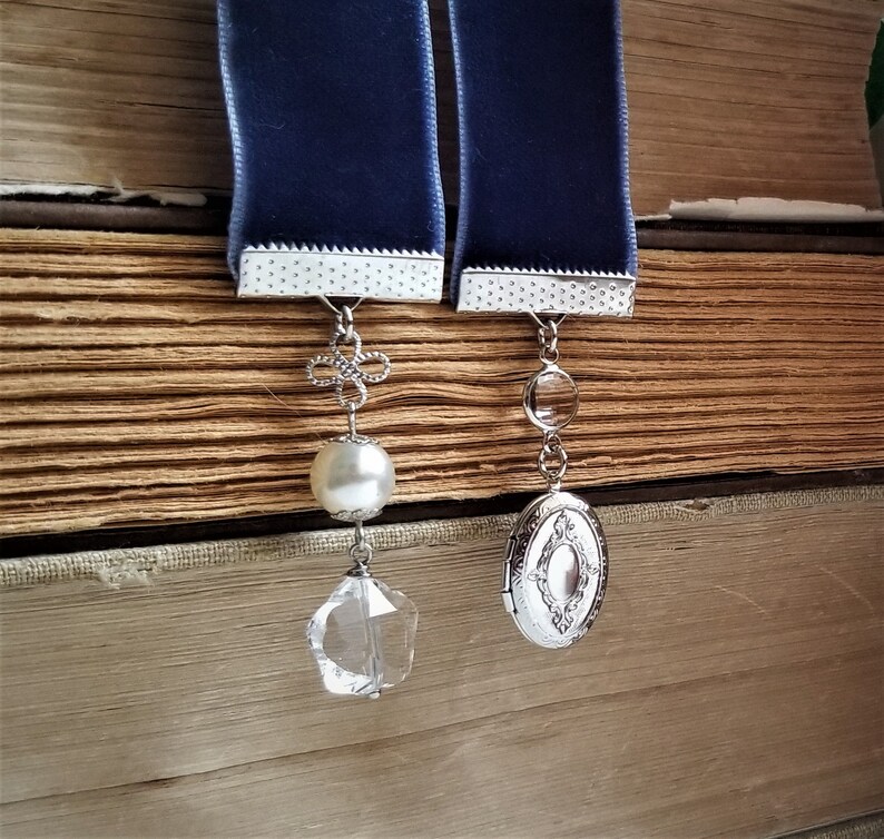 May include: Two blue velvet ribbon bookmarks with silver metal accents. One bookmark has a pearl and a crystal charm, the other has a silver locket charm.