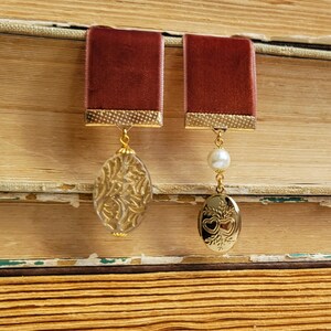 May include: Two burgundy velvet bookmark pendants with gold accents. One pendant features a clear glass charm with a gold floral design. The other pendant features a gold locket with a heart design and a white pearl.