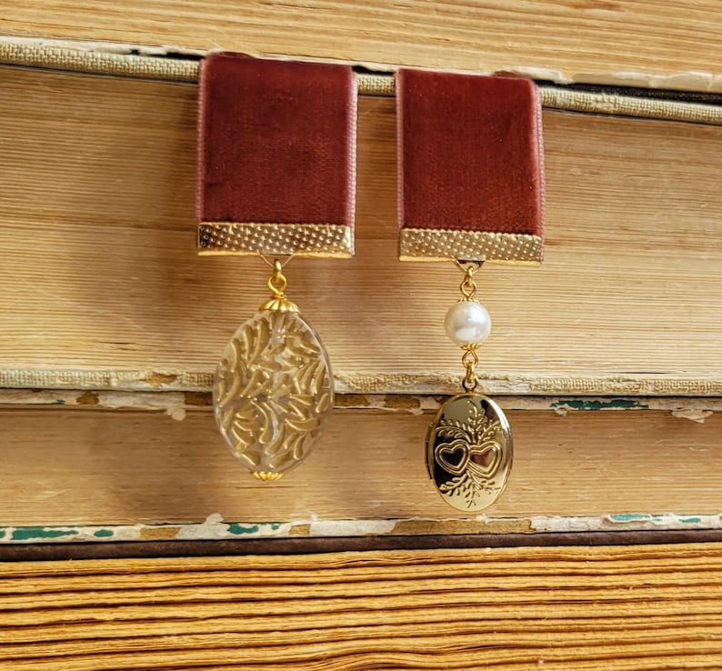 May include: Two burgundy velvet bookmark pendants with gold accents. One pendant features a clear glass charm with a gold floral design. The other pendant features a gold locket with a heart design and a white pearl.
