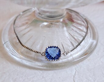 Peut inclure: Un bracelet en argent avec une pierre précieuse carrée bleu clair au centre. La pierre précieuse est sertie dans un cadre argenté décoratif. Le bracelet est présenté sur une surface en verre transparent.