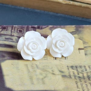 Może przedstawiać: Para białych ceramicznych kolczyków w kształcie róży. Róże są rzeźbione z detalicznym wzorem i mają gładkie, matowe wykończenie.