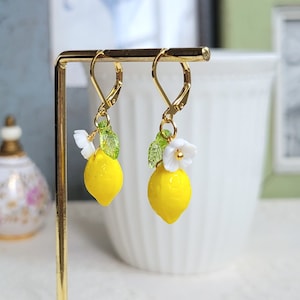 Puede incluir: Pendientes de oro con un dije de limón amarillo, hojas verdes y una flor blanca.