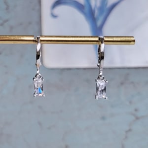 May include: A pair of silver hoop earrings with a dangling rectangular clear stone.