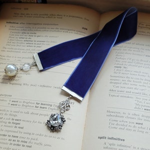 May include: A blue velvet bookmark with a silver charm featuring a teardrop-shaped crystal. The bookmark is lying on an open book with text about grammar and the infinitive.