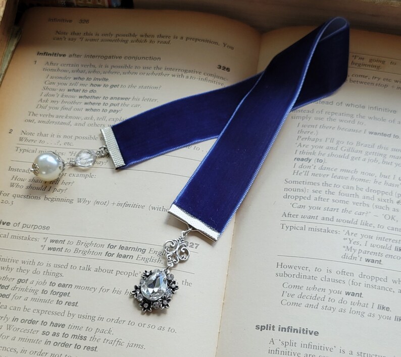 May include: A blue velvet bookmark with a silver charm featuring a teardrop-shaped crystal. The bookmark is lying on an open book with text about grammar and the infinitive.