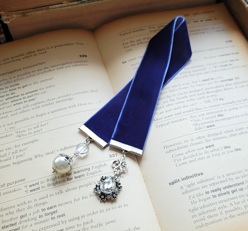 May include: A blue velvet bookmark with a silver charm and a pearl bead. The bookmark is lying open on a book with text about English grammar.