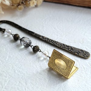 Puede incluir: Un marcapáginas dorado con un pequeño amuleto de libro con bisagras ornamentado y una cadena de cuentas de cristal transparentes. El marcapáginas está hecho de metal y tiene un diseño floral.