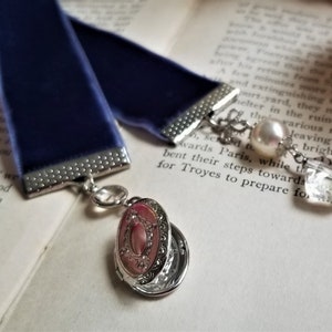 May include: A blue velvet ribbon bookmark with a silver charm featuring a locket and a crystal bead. The bookmark is on a page of a book with text.