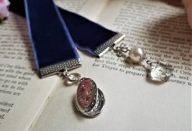 May include: A blue velvet ribbon bookmark with a silver charm featuring a locket and a crystal bead. The bookmark is on a page of a book with text.