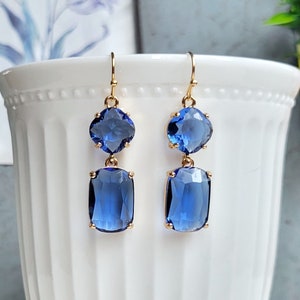 May include: A pair of gold dangle earrings with two blue gemstone drops. The top gemstone is round and the bottom gemstone is rectangular.