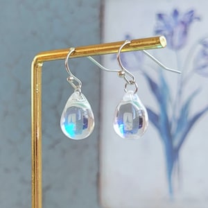 May include: A pair of iridescent teardrop earrings with silver hooks. The earrings are hanging from a gold stand.