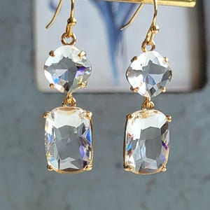 May include: A pair of gold dangle earrings with two clear crystal stones on each earring. The top stone is round and the bottom stone is rectangular.