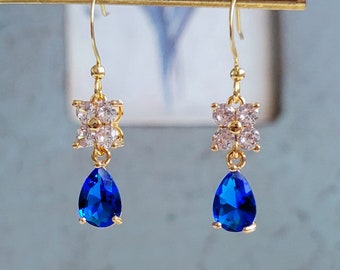 May include: Gold earrings with a blue teardrop-shaped gemstone and a flower-shaped design with clear rhinestones.