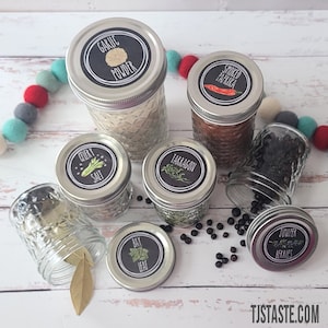 Könnte beinhalten: Eine Sammlung von Glasbehältern mit schwarzen und weißen Etiketten für verschiedene Gewürze und Kräuter. Die Etiketten beinhalten "Knoblauchpulver", "Geräucherter Paprika", "Selleriesalz", "Estragon", "Lorbeerblatt" und "Wacholderbeeren".