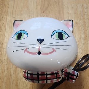 May include: A white ceramic cat head with blue eyes, black whiskers, and a pink smile. The cat wears a red and black plaid scarf and holds a pair of black scissors. The cat is a decorative item.