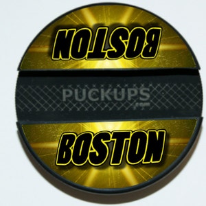May include: A black and yellow hockey puck with the words "BOSTON" and "NOLSON" on it. The puck also has the words "PUCKUPS.COM" on it.