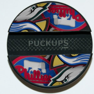 May include: A black and white hockey puck phone grip with a colorful collage of Philadelphia sports team logos, including the Eagles, Phillies, and Flyers. The text "PUCKUPS" is printed on the grip.