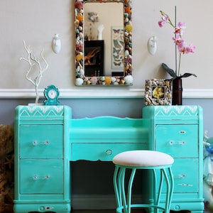 May include: A turquoise vanity set with a mirror framed with seashells, two drawers, a stool, and a small picture frame. The vanity has a white and turquoise chevron pattern on the top.