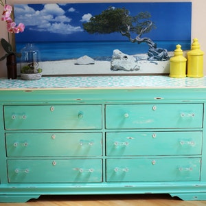 May include: A turquoise dresser with six drawers and clear glass knobs. The dresser has a distressed paint finish and a white top with a stenciled design. Two yellow ceramic jars are on top of the dresser.