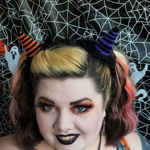 May include: A woman wearing a black and orange Halloween-themed dress with a black cat pumpkin and a necklace with two pumpkin-shaped charms. She has black eyeliner and orange eyeshadow.
