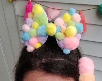 May include: A pastel pink, yellow, and blue pom-pom bow headband. The headband is worn by a person with dark hair and pink eyeshadow.