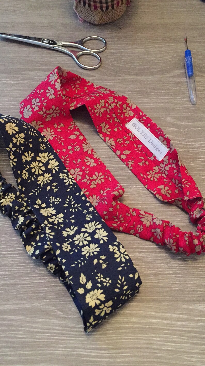 May include: Two floral headbands, one red and one navy, are displayed on a wooden surface. The red headband features a beige floral pattern, while the navy headband has a similar beige floral design. The headbands are elasticated.