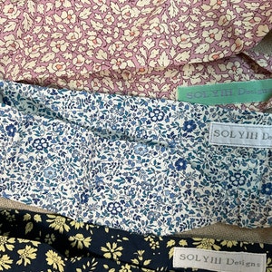 May include: Three floral headbands in shades of pink, blue, and navy. Each headband features a different floral pattern and a tag that reads "SOLYIH Designs". The headbands are made of a soft fabric.