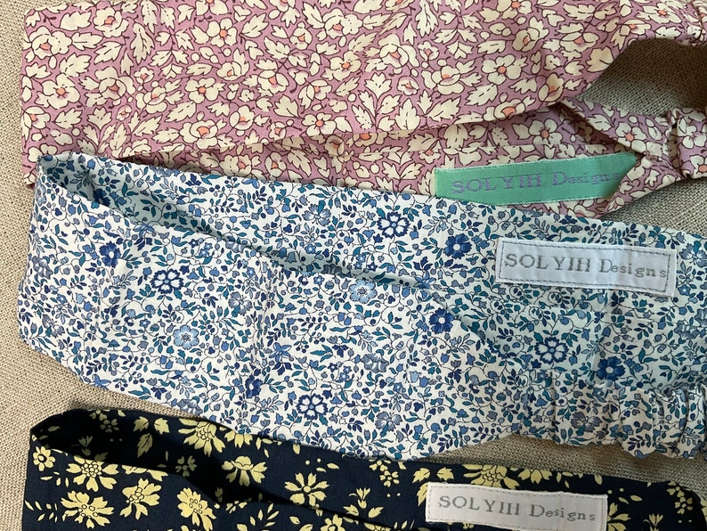 May include: Three floral headbands in shades of pink, blue, and navy. Each headband features a different floral pattern and a tag that reads "SOLYIH Designs". The headbands are made of a soft fabric.