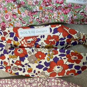 May include: Close-up of several floral headbands. The headbands feature a variety of colorful floral patterns, including red, orange, purple, and pink flowers on a cream background. The label "SOLYIII Designs" is visible on one headband.