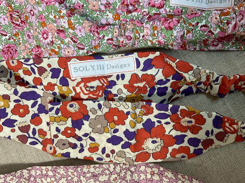 May include: Close-up of several floral headbands. The headbands feature a variety of colorful floral patterns, including red, orange, purple, and pink flowers on a cream background. The label "SOLYIII Designs" is visible on one headband.