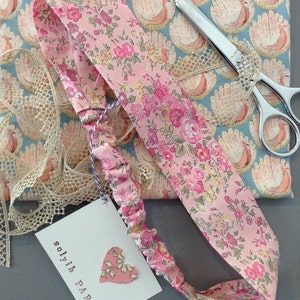 May include: A pink floral headband with a matching tag that reads "solyih PARIS". The headband is made of a soft, patterned fabric with a delicate floral design. A pair of scissors and lace trim are also visible.