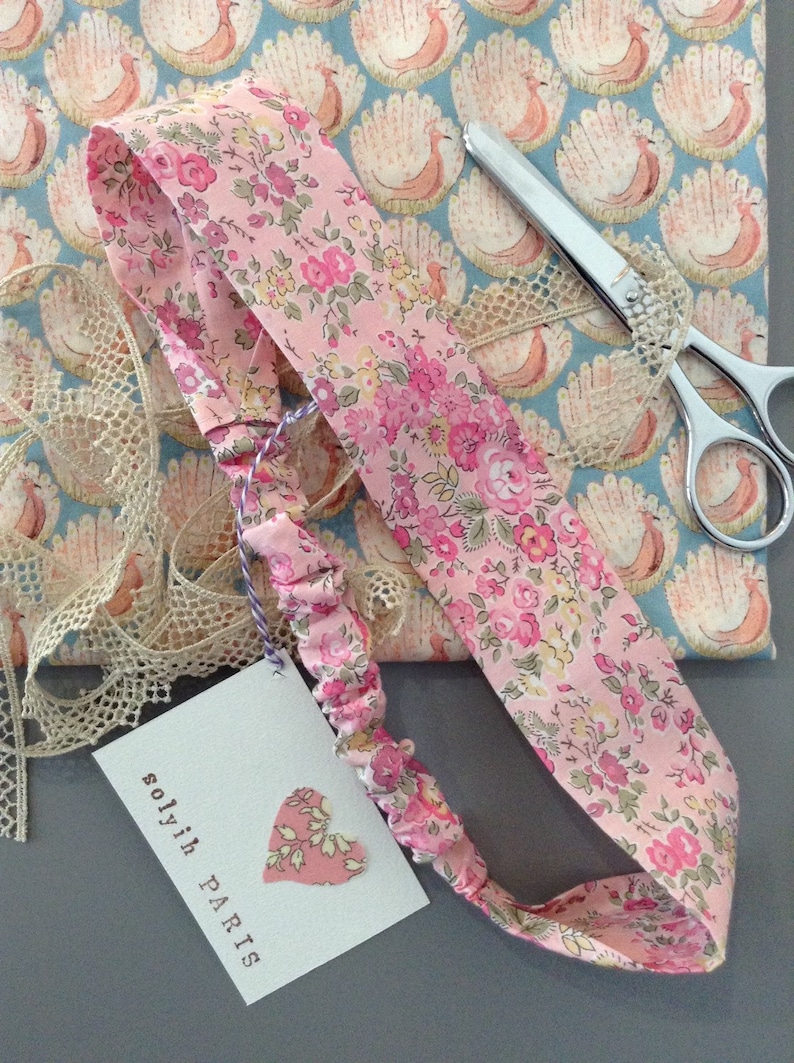 May include: A pink floral headband with a matching tag that reads "solyih PARIS". The headband is made of a soft, patterned fabric with a delicate floral design. A pair of scissors and lace trim are also visible.