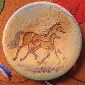 May include: A round ceramic dish with a speckled beige and green glaze featuring a raised, embossed design of two horses running. The surface has a textured, earthy appearance with brown flecks throughout the glaze, highlighting the equine figures.