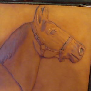 May include: A brown ceramic tile with a relief sculpture of a horse's head. The horse is wearing a leather bridle with a metal buckle.